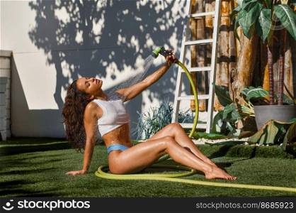 Beautiful young woman refreshing herself from garden hose sitting on backyard. Hot summer day. Young woman refreshing herself from garden hose