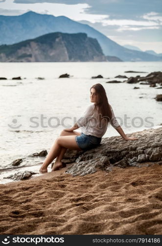 Beautiful young woman relaxing sitting on big rock on beach and looking at far away island