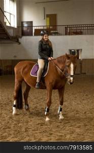 Beautiful young woman riding brown horse in indoor manege