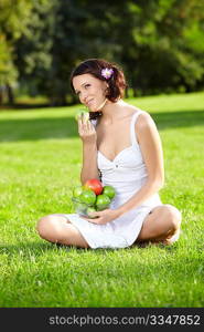 Beautiful young woman sits and eats an apple on lawn in summer garden