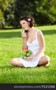 Beautiful young woman sits and eats an apple on lawn in summer garden