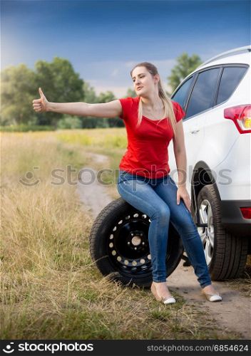 Beautiful young woman sitting at broken car and hitchhiking