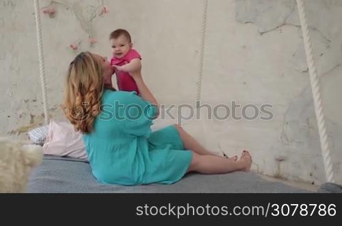 Beautiful young woman sitting on bed in stylish bedroom, bonding with her adorable baby girl while family spending leisure at home. Loving mother gently kissing her infant child. Slow motion. Steadicam stabilized shot.