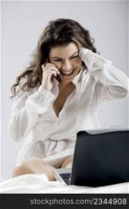Beautiful young woman sitting on bed working with a laptop and making a phone call