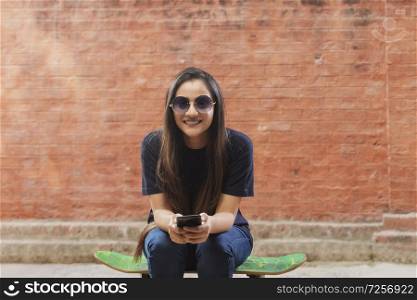 Beautiful young woman sitting on skateboard on Street Wearing Sunglasses, using smart phone
