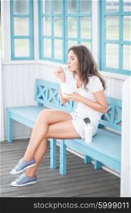 Beautiful young woman, sitting on the bench, holding a fast food lunch box, eating up noodles from Chinese take-out with chopsticks and drinking takeaway coffee.