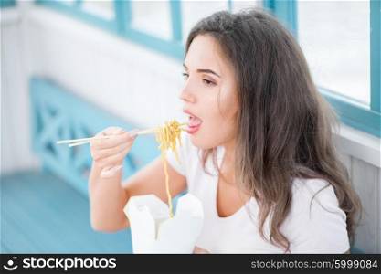 Beautiful young woman, sitting on the bench, holding a fast food lunch box and eating up noodles from Chinese take-out with chopsticks.