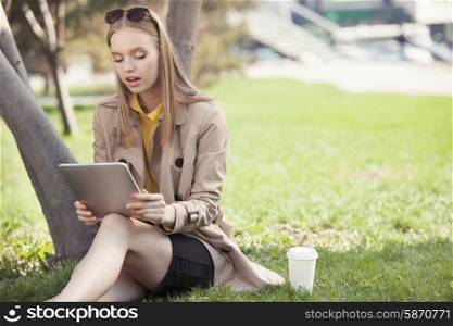 Beautiful young woman sitting on the grass with a disposable coffee cup, holding tablet in her hands, reading and studying against summer park background.