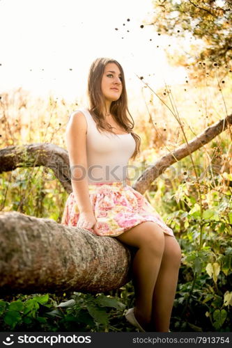Beautiful young woman sitting on tree branch at field at sunny day