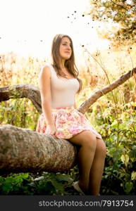 Beautiful young woman sitting on tree branch at field at sunny day