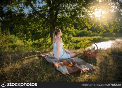 Beautiful young woman sitting under big tree at field and looking at sunset
