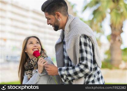 Beautiful young woman smiles as she receives a red rose from a handsome young man on an out of focus background. Valentine?s concept.. Young woman receives a red rose from a young man
