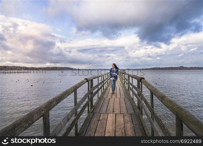 Beautiful young woman smiling and walking alone on a long wooden pontoon, over the lake Chiemsee, enjoying the fresh air and the sunset, in Germany.