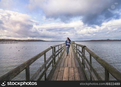 Beautiful young woman smiling and walking alone on a long wooden pontoon, over the lake Chiemsee, enjoying the fresh air and the sunset, in Germany.