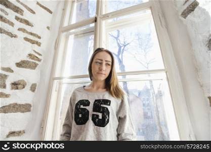 Beautiful young woman standing against window at home