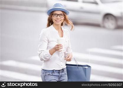 Beautiful young woman standing at the crosswalk with a coffee-to-go cup, smiling happily against urban city background.