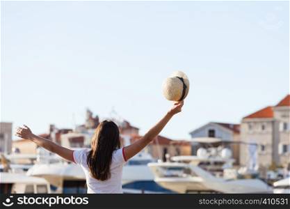 Beautiful young woman standing close to the boats at Mediterranean city marina port and holding hat