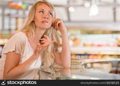 Beautiful young woman standing near supermarket showcase full of different products and deciding what to buy