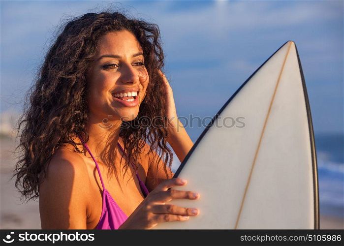 Beautiful young woman surfer girl in bikini with surfboard standing on a beach at sunset or sunrise