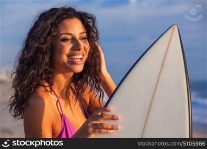 Beautiful young woman surfer girl in bikini with surfboard standing on a beach at sunset or sunrise