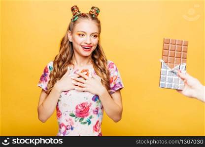 Beautiful young woman surprised somebody gives her big chocolate bar. Bright girl with blonde curly hair. Stylish girl in summer colorful dress, yellow wall on background.. Woman surprised somebody gives her big chocolate
