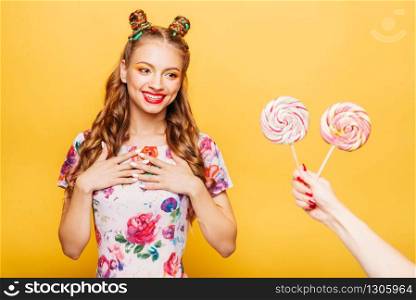 Beautiful young woman surprised somebody gives her lollypops. Bright girl with blonde curly hair. Stylish girl in summer colorful dress, yellow wall on background.. Woman surprised somebody gives her lollypops