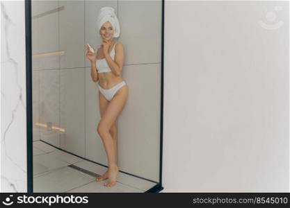 Beautiful young woman using cosmetic product for healthy skin and smiling while posing in bathroom at home, doing beauty routine after morning shower, wears wrapped towel on head and white lingerie. Beautiful young woman using cosmetic product for healthy skin and smiling while posing in bathroom at home