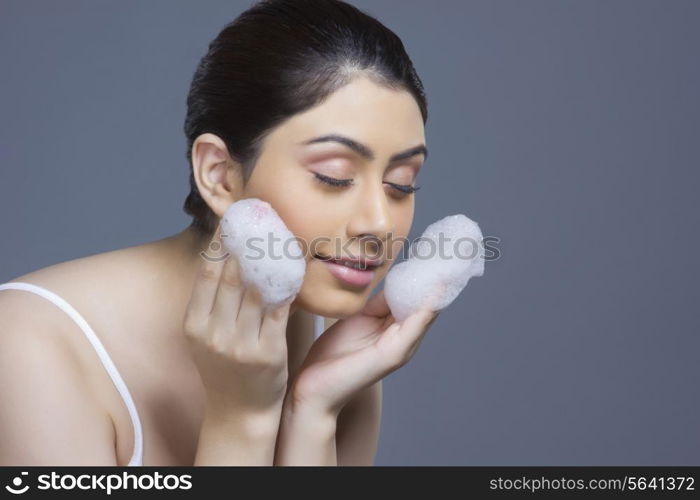 Beautiful young woman washing face with soap sud over blue background