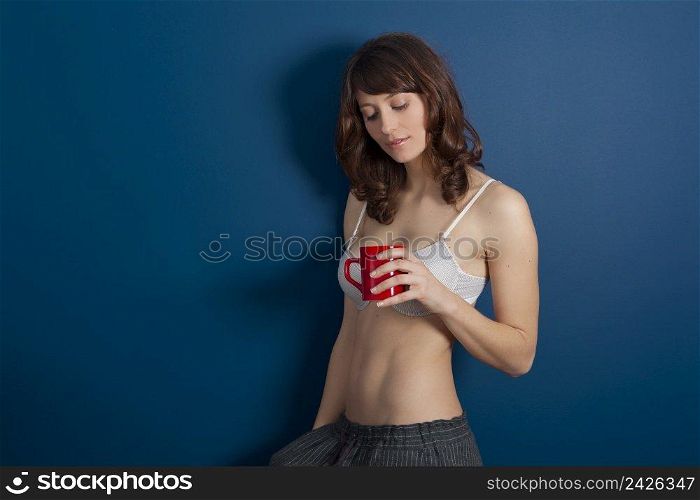 Beautiful young woman wearing a casual lingerie against a blue wall, and drinking coffee