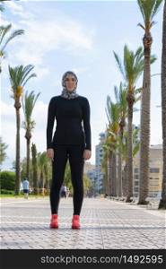 Beautiful young woman wearing a headscarf, tights and a long sleeve sports shirt taking a break in the middle of the street on an out of focus background. Diversity and healthy lifestyle concept.
