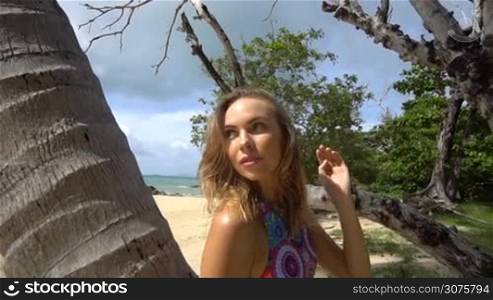 Beautiful young woman wearing colorful bikini on a beautiful summer day standing, leaning against a palm tree - video in slow motion