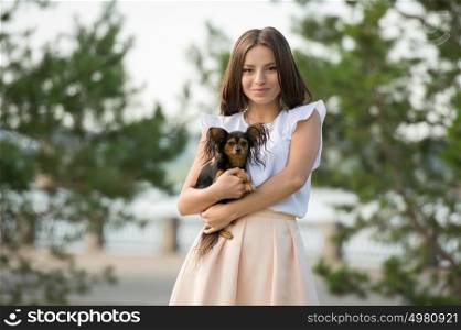 Beautiful young woman wearing elegant dress holding small dog while walking in summer park