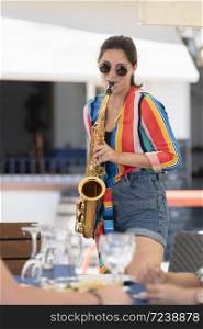 Beautiful young woman wearing glasses and playing a saxophone close to an out of focus outdoor bar terrace on an out of focus background. Street performer and lifestyle concept.. young woman wearing glasses and playing a saxophone close to an out of focus outdoor bar terrace