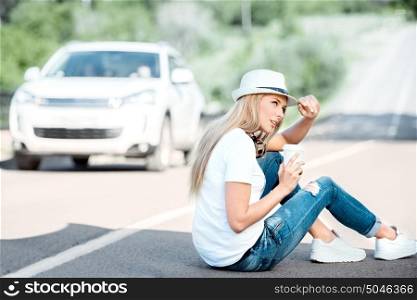 Beautiful young woman wearing hat and music headphones around her neck, drinking coffee from a takeaway coffee cup and sitting on a separating strip on the road.