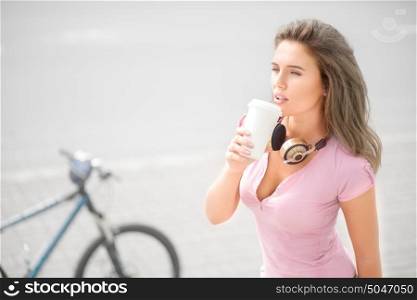 Beautiful young woman wearing music headphones around her neck, drinking coffee from a take away coffee cup against bicycle and road background.
