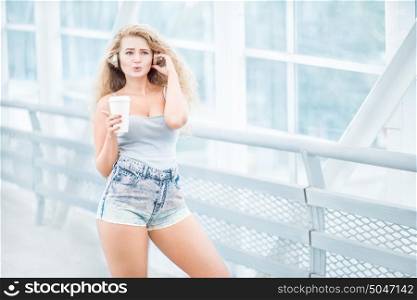 Beautiful young woman wearing music headphones, standing on the bridge with a take away coffee cup and posing against urban background.