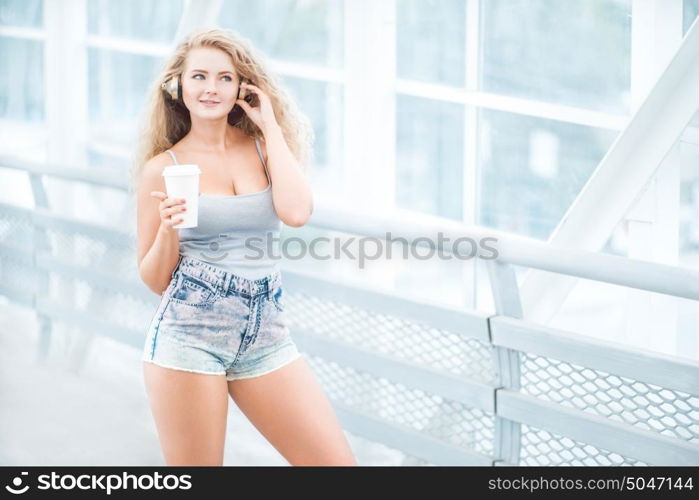 Beautiful young woman wearing music headphones, standing on the bridge with a take away coffee cup and posing against urban background.