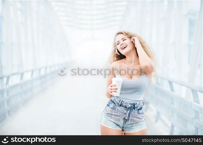 Beautiful young woman wearing music headphones, standing on the bridge with a take away coffee cup and posing against urban background.