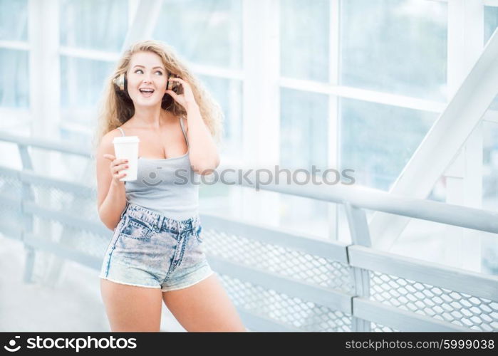 Beautiful young woman wearing music headphones, standing on the bridge with a take away coffee cup and posing against urban background.