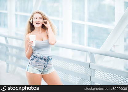 Beautiful young woman wearing music headphones, standing on the bridge with a take away coffee cup and posing against urban background.