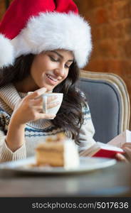 Beautiful young woman wearing Santa Claus red hat sitting at cafe and having hot beverage and tasty cake while reading a book