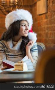 Beautiful young woman wearing Santa Claus red hat sitting at cafe and having hot beverage and tasty cake while reading a book