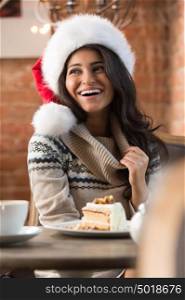 Beautiful young woman wearing Santa Claus red hat sitting at cafe and having hot beverage and tasty cake