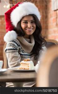 Beautiful young woman wearing Santa Claus red hat sitting at cafe and having hot beverage and tasty cake