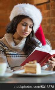 Beautiful young woman wearing Santa Claus red hat sitting at cafe and having hot beverage and tasty cake while reading a book