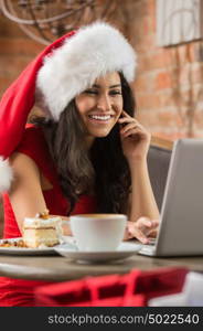 Beautiful young woman wearing Santa Claus red hat sitting at cafe and having hot beverage and tasty cake while using her laptop