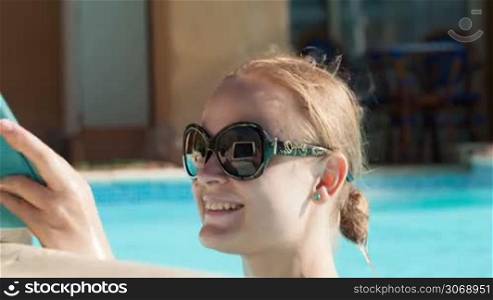 Beautiful young woman wearing sunglasses using a tablet poolside resting her arms on the pool surround as she reads information on the screen