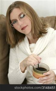 Beautiful young woman wearing sweater and glasses. Sitting on couch with cup of coffee.