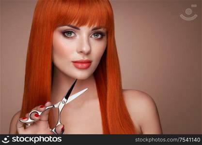 Beautiful young woman with a bright makeup and a smooth long hair holds metal scissors. Model with red hair. Hair salon, haircut. Care and beauty hair products. Perfect make-up