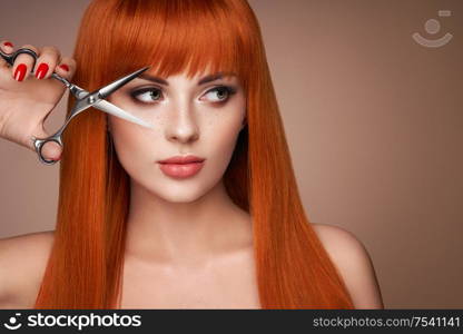 Beautiful young woman with a bright makeup and a smooth long hair holds metal scissors. Model with red hair. Hair salon, haircut. Care and beauty hair products. Perfect make-up